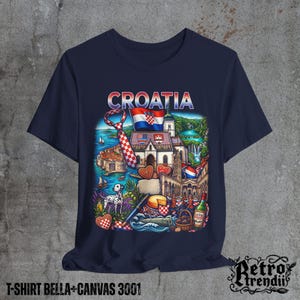 A blue T-shirt featuring a colorful graphic design of Croatia landmarks and symbols, including a Dalmatian dog, necktie, Mediterranean coast, and historic architecture, displayed on a textured grey background.