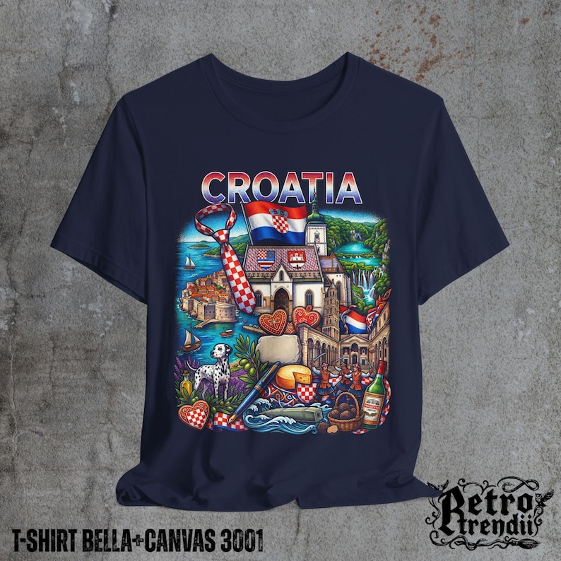 A blue T-shirt featuring a colorful graphic design of Croatia landmarks and symbols, including a Dalmatian dog, necktie, Mediterranean coast, and historic architecture, displayed on a textured grey background.