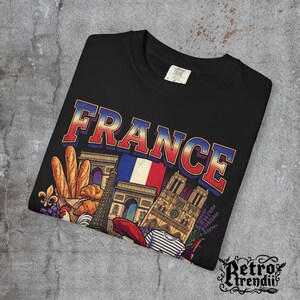 A black acid-wash T-shirt featuring a colorful graphic design of France landmarks and symbols, including the Eiffel Tower, Notre-Dame, croissants, wine, and French fashion, showcasing European culture.