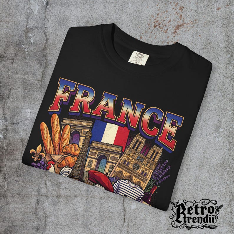 A black acid-wash T-shirt featuring a colorful graphic design of France landmarks and symbols, including the Eiffel Tower, Notre-Dame, croissants, wine, and French fashion, showcasing European culture.