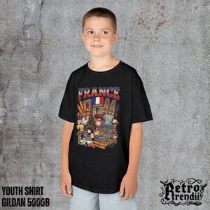A young boy wearing a black T-shirt featuring a colorful graphic design of France landmarks and symbols, including the Eiffel Tower, Notre-Dame, croissants, wine, and French fashion.