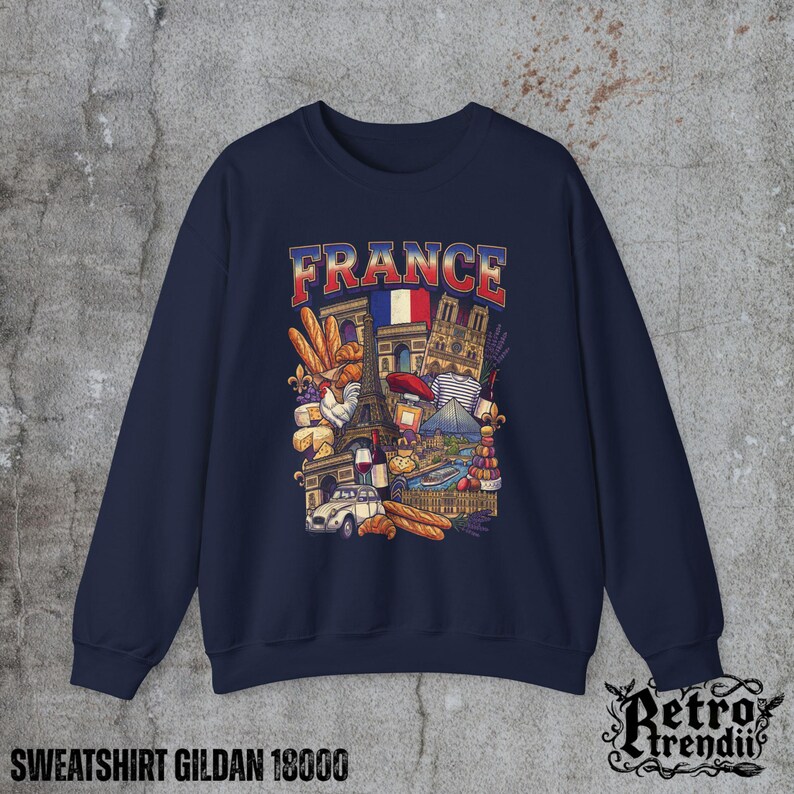 A blue crewneck sweatshirt featuring a detailed graphic design of France landmarks and symbols, including the Eiffel Tower, Notre-Dame, croissants, wine, and French fashion, displayed on a textured grey background.