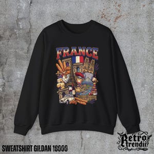 A black crewneck sweatshirt featuring a detailed graphic design of France landmarks and symbols, including the Eiffel Tower, Notre-Dame, croissants, wine, and French fashion, displayed on a textured grey background.