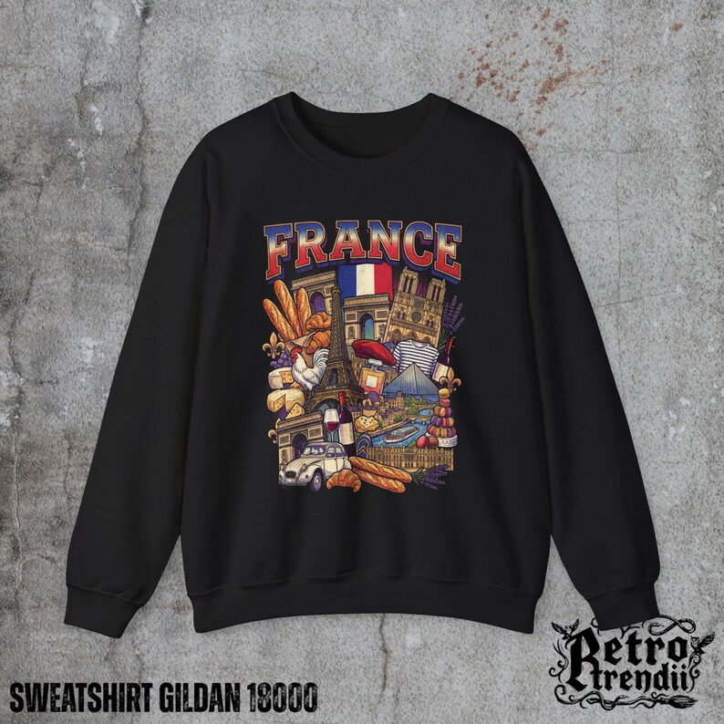 A black crewneck sweatshirt featuring a detailed graphic design of France landmarks and symbols, including the Eiffel Tower, Notre-Dame, croissants, wine, and French fashion, displayed on a textured grey background.
