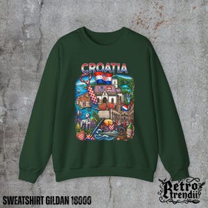 A Green crewneck sweatshirt featuring a detailed graphic design of Croatia landmarks and symbols, including a Dalmatian dog, necktie, Mediterranean coast, and historic architecture, displayed on a textured grey background.
