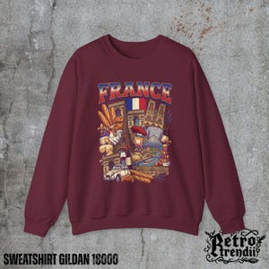 A Red crewneck sweatshirt featuring a detailed graphic design of France landmarks and symbols, including the Eiffel Tower, Notre-Dame, croissants, wine, and French fashion, displayed on a textured grey background.