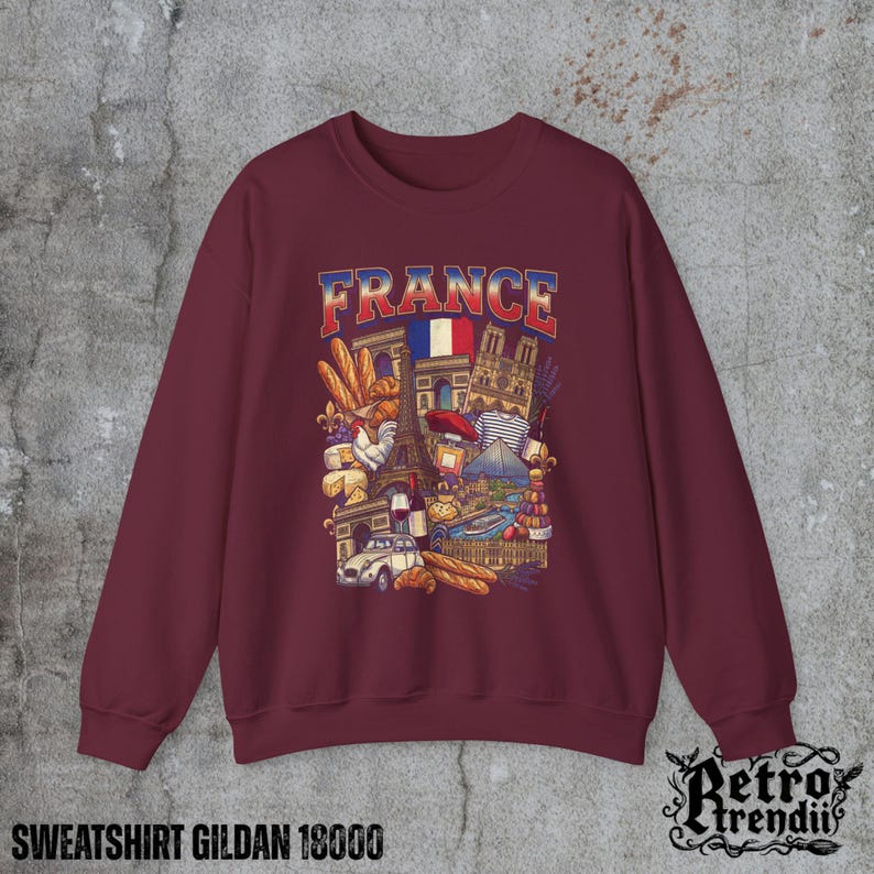 A Red crewneck sweatshirt featuring a detailed graphic design of France landmarks and symbols, including the Eiffel Tower, Notre-Dame, croissants, wine, and French fashion, displayed on a textured grey background.
