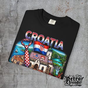 A black acid-wash T-shirt featuring a colorful graphic design of Croatia landmarks and symbols, including a Dalmatian dog, necktie, Mediterranean coast, and historic architecture, showcasing European culture.