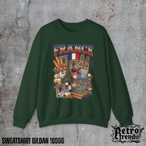 A Green crewneck sweatshirt featuring a detailed graphic design of France landmarks and symbols, including the Eiffel Tower, Notre-Dame, croissants, wine, and French fashion, displayed on a textured grey background.