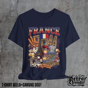 A blue T-shirt featuring a colorful graphic design of France landmarks and symbols, including the Eiffel Tower, Notre-Dame, croissants, wine, and French fashion, displayed on a textured grey background.