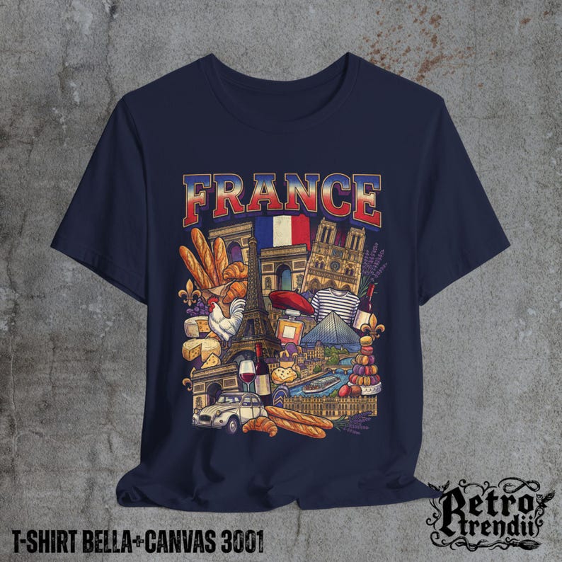 A blue T-shirt featuring a colorful graphic design of France landmarks and symbols, including the Eiffel Tower, Notre-Dame, croissants, wine, and French fashion, displayed on a textured grey background.