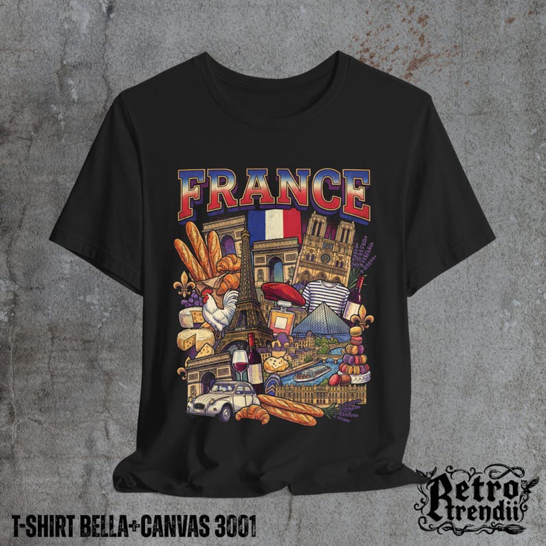 A black T-shirt featuring a colorful graphic design of France landmarks and symbols, including the Eiffel Tower, Notre-Dame, croissants, wine, and French fashion, displayed on a textured grey background.