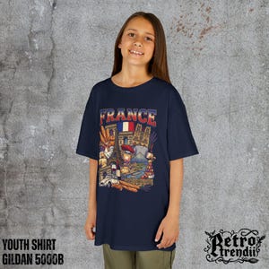 A young girl wearing a blue T-shirt featuring a colorful graphic design of France landmarks and symbols, including the Eiffel Tower, Notre-Dame, croissants, wine, and French fashion.