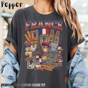 A person wearing a grey pepper acid-wash T-shirt featuring a colorful graphic design of France landmarks and symbols, including the Eiffel Tower, Notre-Dame, croissants, wine, and French fashion, showcasing European culture.
