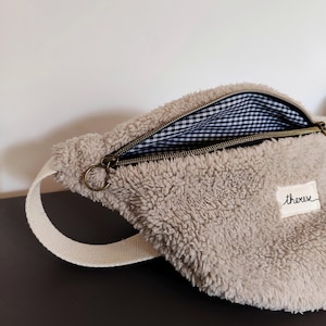 Peut inclure: Un sac banane beige et duveteux avec une sangle crème. L'intérieur est doublé d'un motif vichy noir et blanc. La fermeture éclair est ouverte et le mot "thereve" est brodé sur un petit patch blanc.