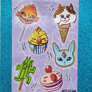 Könnte beinhalten: Ein Bogen mit Aufklebern mit Cartoon-Essen und Charakterdesigns. Enthält einen Lolli, eine Eistüte, einen Cupcake, einen Keks, einen Eisriegel und ein Macaron. Die Aufkleber sind in verschiedenen Farben gehalten, darunter Orange, Braun, Gelb, Blau und Grün. Der Text "KITCATLAW" steht unten.