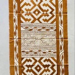Könnte beinhalten: Ein rechteckiger Stoffläufer mit einem markanten, sich wiederholenden geometrischen Muster in tiefem Ockerbraun und Off-White. Das Design umfasst verschlungene, winklige Linien, ein zentrales Band mit feinen linearen Motiven und eine Bordüre aus stilisierten Blattformen.