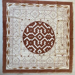 May include: A square fabric textile featuring a central brown circle with a white geometric maze pattern. The surrounding square border displays a repeating brown and white linear design, including zigzags and leaf-like shapes on the outer edge.