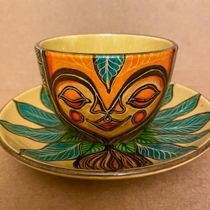 May include: A ceramic teacup and saucer set. The cup and saucer are yellow with a painted face design in orange, black, and red. The saucer has blue leaf accents. The cup has a matching face design.
