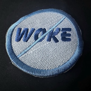 May include: Embroidered patch with a light blue border and a light gray center. The word "WOKE" is embroidered in dark blue, with a blue diagonal line through it. The patch has a slightly irregular, rounded shape.
