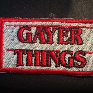 May include: Embroidered patch with the words "GAYER THINGS" in red, stylized lettering. The text is on a light gray background, framed by a red border. The patch is rectangular and appears to be iron-on.