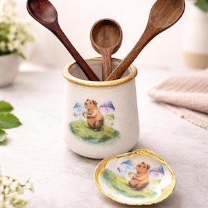 May include: A white ceramic utensil holder with a gold rim, featuring a whimsical illustration of a bear. Three wooden spoons of varying sizes are inside. A matching seashell-shaped dish is in front.