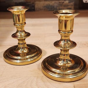 May include: A pair of polished gold-tone metal candle holders featuring a wide circular base, a slender central stem with decorative tiered rings, and a flared top opening designed to securely hold a standard taper candle in an upright position.