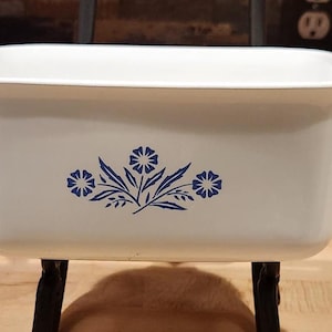 May include: A rectangular white ceramic baking dish with integrated side handles. The front of the dish features a centered blue floral pattern consisting of three stylized flowers with pointed leaves and stems arranged in a symmetrical design.