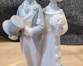 May include: A porcelain figurine of a bride and groom in traditional wedding attire. The bride is wearing a white dress with a lace trim and a floral crown. The groom is wearing a gray suit with a white shirt and a top hat. The figurine is a traditional wedding decoration.