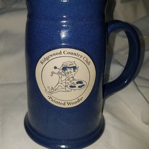 May include: Blue ceramic beer stein with a white circle logo that says "Ridgewood Country Club" and features a cartoon elf holding a paintbrush and palette. The text "Jack Frost" and "Painted Woods" are also on the logo.