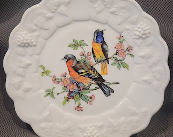 May include: White milk glass plate with a raised embossed design of leaves and grapes around the edge. The center features a painted image of two birds perched on a branch with pink flowers.