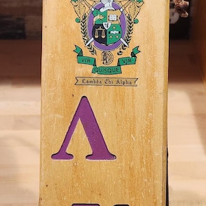 May include: A light-colored wooden paddle featuring the Lambda Chi Alpha fraternity crest in green, purple, and gold. The Greek letters Lambda, Chi, and Alpha are printed in purple below the crest. The text FALL WEEK END 1956 is printed in black.
