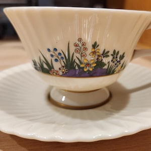 May include: Cream-colored teacup and saucer set. The teacup features a floral design with blue, yellow, and red flowers, green leaves, and a purple base. The saucer has a ridged, scalloped edge and a matching cream color.