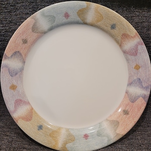 May include: A white ceramic dinner plate with a colorful abstract design in shades of pink, blue, and yellow. The design features a series of overlapping shapes and swirls.