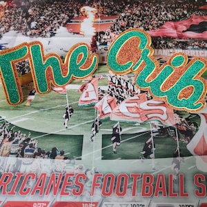 Könnte beinhalten: Zwei grüne und goldene Glitzer-Schilder, auf denen in kursiv "The Crib" steht. Die Schilder befinden sich vor einem Hintergrund eines Fußballspiels.