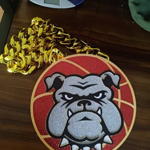 Könnte beinhalten: Eine goldene Kettenkette mit einem runden roten und weißen Anhänger, der einen Cartoon-Bulldog mit einem schwarzen Halsband und weißen Spikes zeigt. Der Bulldog hat ein weißes Gesicht mit schwarzen Augen und einer schwarzen Nase.