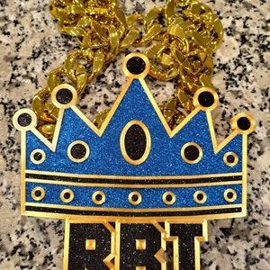 Könnte beinhalten: Eine goldene Kettenkette mit einem blauen und schwarzen Kronenanhänger, auf dem "RBI" in schwarzem Glitzer steht.