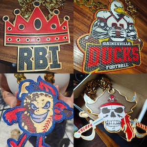 Könnte beinhalten: Vier verschiedene goldene Ketten mit Anhängern. Der erste Anhänger ist eine rote Krone mit den Buchstaben RBI in Gold. Der zweite Anhänger ist ein rot-schwarzer Fußballspieler mit dem Text "Gainesville Ducks Football". Der dritte Anhänger ist eine blaue und rote Comicfigur mit einem Baseball-Design. Der vierte Anhänger ist ein weißer Schädel mit einem roten Bandana und zwei gekreuzten Schwertern.