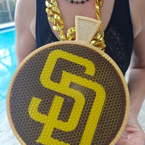 Könnte beinhalten: Ein großer goldener Anhänger mit einem Wabenmuster und dem San Diego Padres Logo in Gelb. Der Anhänger hängt an einer dicken goldenen Kette.