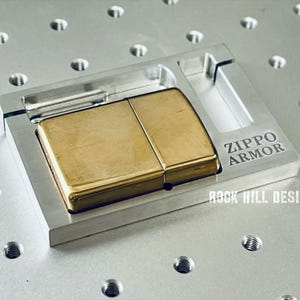 May include: A gold-colored Zippo lighter in a silver metal holder. The holder has the text "ZIPPO ARMOR" and "ROCK HILL DESIGNS" engraved on it. The lighter is rectangular and has a shiny finish.