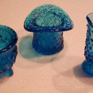 Könnte beinhalten: Drei blaue Glasstücke: eine kleine Schale mit drei Beinen, eine schalenförmige Mütze und ein Krug mit Henkel. Alle drei Stücke haben eine strukturierte, unebene Oberfläche.