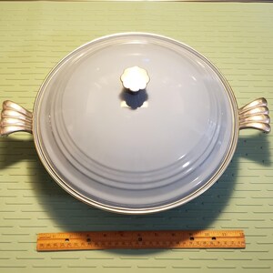 May include: A round, light blue casserole dish with a clear glass lid and silver-colored handles. The lid has a flower-shaped knob. The dish is approximately 12 inches in diameter.