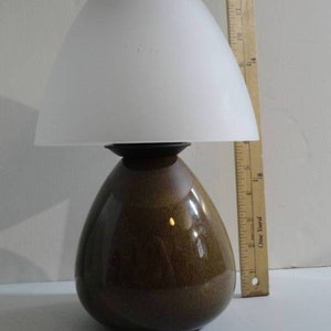 Puede incluir: Una lámpara de mesa de cerámica marrón con una pantalla de vidrio blanco esmerilado. La lámpara tiene forma de lágrima y un acabado liso y brillante.