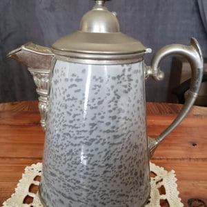 Könnte beinhalten: Eine silberne Kanne mit einem gesprenkelten grauen Emaille-Finish und einem dekorativen Griff. Die Kanne hat einen Deckel mit einem Knauf oben.