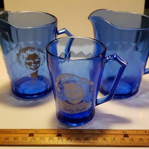 Puede incluir: Tres piezas de cristalería azul Shirley Temple. El juego incluye dos jarras y una taza, cada una con una imagen plateada de Shirley Temple. La taza tiene un asa geométrica y el nombre "Shirley Temple" impreso.