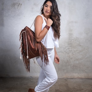 May include: A brown leather backpack with long fringe detail and white rope straps. The backpack is carried over the shoulder. The person is wearing a white top, white pants, and brown suede shoes. A brown leather bracelet is on the wrist.