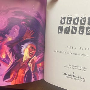 May include: A book titled "Dead Lines" by Greg Bear with a purple and red illustration of a man on the cover. The book is a signed first edition bound in genuine leather and published by The Easton Press.