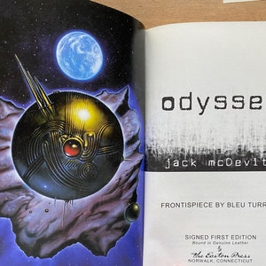 May include: A book cover with a dark blue background and a silver spaceship in the foreground. The title "odyssey" is written in black text on the cover, with the author's name "jack mcDevitt" written below it in smaller text. The cover also includes the text "FRONTISPIECE BY BLEU TURRELL" and "SIGNED FIRST EDITION Bound in Genuine Leather The Easton Press NORWALK, CONNECTICUT".