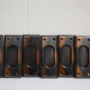 May include: Six rectangular, bronze-colored recessed pulls with oval finger holes. The pulls have a dark, matte finish with copper-colored accents. Each pull has two screw holes at the top and bottom.