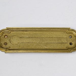 May include: A gold-toned metal mail slot with a decorative border and two small holes. The slot is rectangular with a rounded opening.